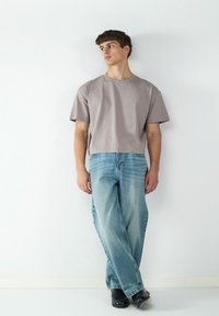 Szary krótki t-shirt o luźnym kroju, zestawiony z jasnoniebieskimi, luźnymi dżinsami. Model nosi czarne buty, stoi na jednolitym tle.