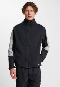 Ung man iklädd en svart zip-up jacka med ljusgrå ränder på ärmarna och matchande svarta byxor, stående mot en vit bakgrund.