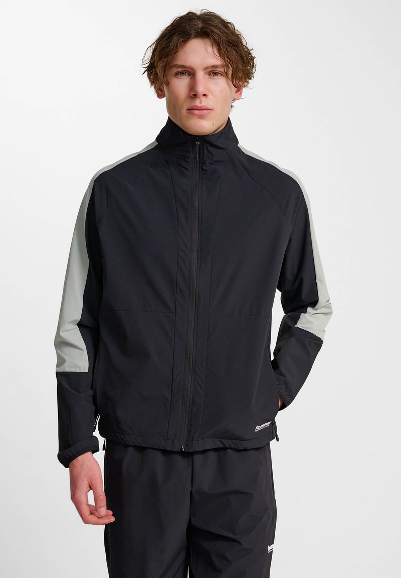 Ung man iklädd en svart zip-up jacka med ljusgrå ränder på ärmarna och matchande svarta byxor, stående mot en vit bakgrund.