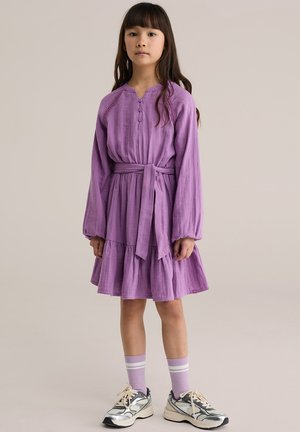 Lila hosszú ujjú ruha réteges szoknyával, elején gombokkal és övvel. Szürke és ezüst sportcipőkkel, valamint csíkos lila zoknival viselve.