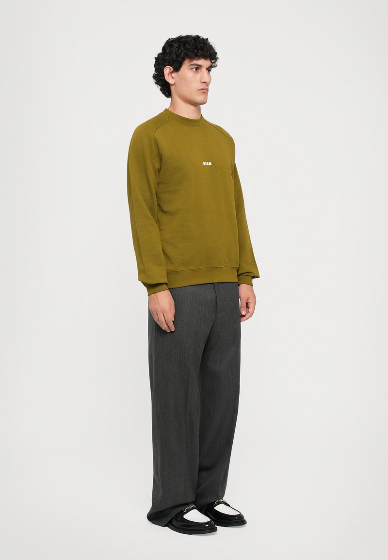 Un homme portant un sweat-shirt vert olive, un pantalon gris ample et des mocassins noir et blanc se tient devant un fond clair.