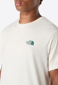 Tæt på en person, der bærer en off-white T-shirt med et lille grønt "The North Face" logo på venstre bryst.