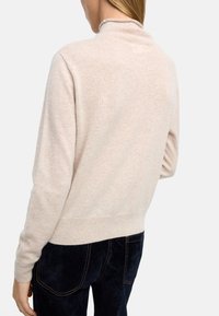 Jasnobeżowy sweter z półgolfem, prążkowanymi mankietami i wykończeniem. Miękka tekstura i dopasowany krój. Noszony z ciemnoniebieskimi spodniami, prezentujący widok z tyłu.