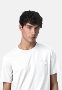 T-shirt in cotone bianco con colletto rotondo, maniche corte e una piccola tasca sul petto sinistro con una toppa del logo. Tessuto liscio.