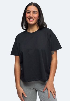 Lächelnde Frau mit dunklen Haaren trägt ein lockeres schwarzes T-Shirt und graue Hosen und steht vor einem schlichten hellen Hintergrund.