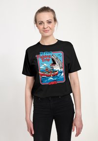 Žena v černém tričku s kresleným motivem dvou lidí lovících ryby v lodi, za nimiž z vody skáče velký žralok.