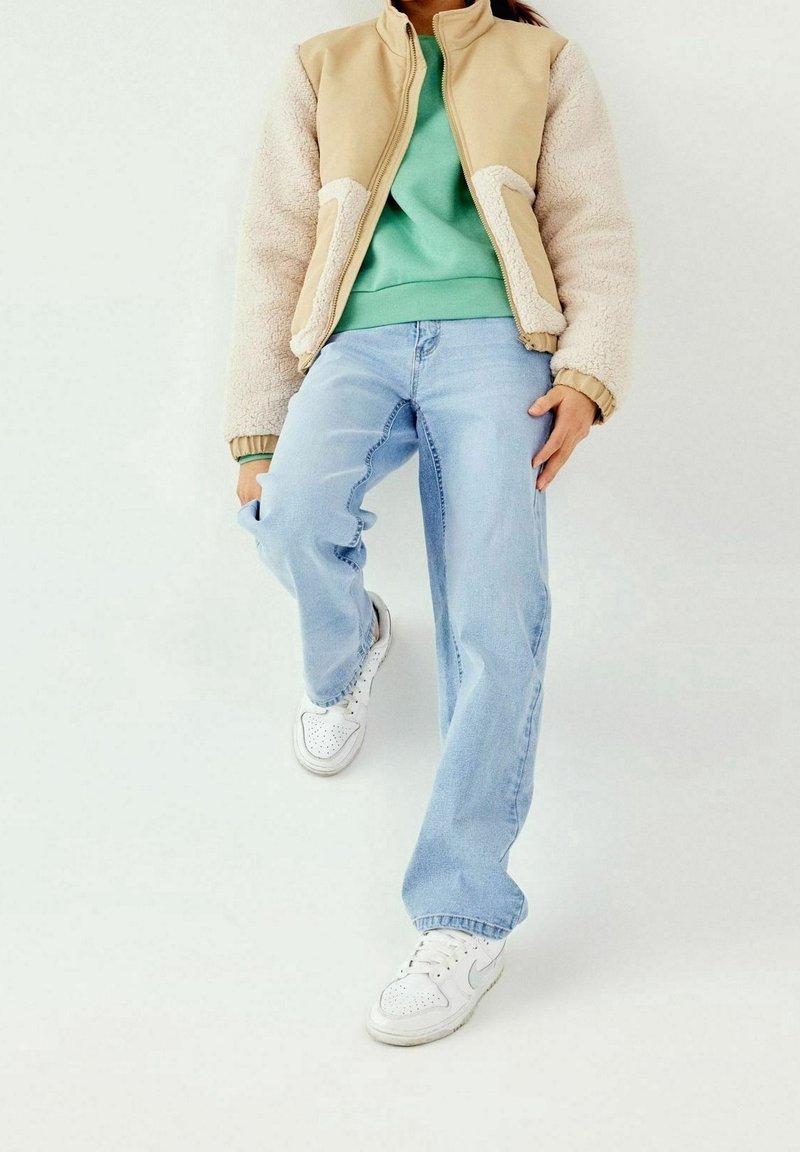 Personne portant une veste en polaire beige, un sweat-shirt vert, un jean bleu clair et des baskets blanches, se tenant avec un genou levé contre un fond uni.