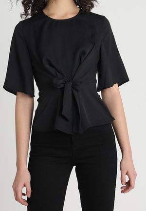 Femme portant un haut noir à manches courtes noué à la taille, associé à un pantalon noir, sur un fond clair uni.
