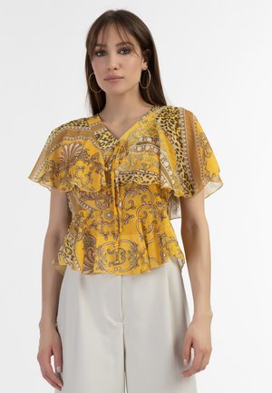 Gul bluse med et lagdelt, flæset design og V-hals. Har indviklede brune mønstre og en snøre foran.