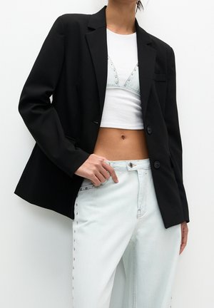 Frau trägt schwarzen Blazer über weißem Croptop und hellblauen, mit Nieten besetzten Jeans vor weißem Hintergrund.