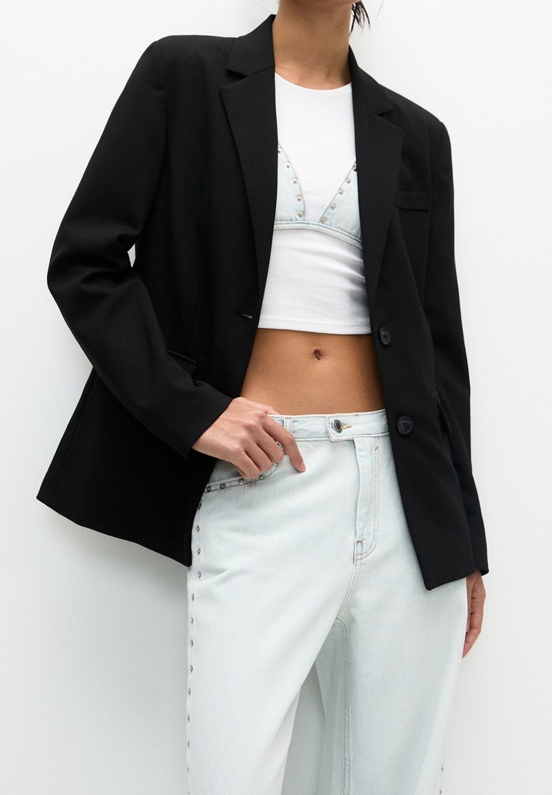 Femme portant un blazer noir sur un crop top blanc et un jean bleu clair clouté sur fond blanc.