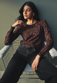 Ilga rankovė viršuje ruda ir juoda leopardų rašto, derinama su aukštu liemeniu juodomis džinsinėmis kelnėmis, sėdint ant metalinės kėdės.