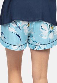 Lagelijf van een persoon in lichtblauwe bloemen shorts met donkere bies en een marineblauwe top, staande tegen een effen achtergrond.