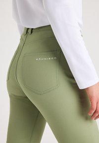 Nahaufnahme einer Person in olivgrünen, enganliegenden Hosen mit Röhnisch-Logo auf der Rückentasche und einem weißen Langarmoberteil, die mit entspanntem Hand steht.