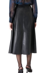 Jupe midi en cuir noir avec une silhouette évasée, texture lisse et détails de couture. Portée avec des chaussures à talons hauts noires et des collants transparents.