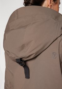 Brown hooded jacket with textured fabric. Features a black strap and round metal hardware detail. Logo printed on the side in gray.