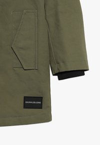 Kurtka oliwkowa z dużą klapą na kieszeń i czarnymi ściągaczami przy mankietach. Zawiera czarną metkę z logo "Calvin Klein Jeans".