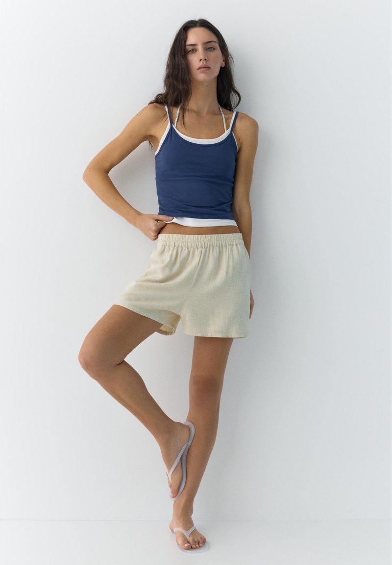 Jeune femme debout contre un mur blanc, portant un débardeur bleu marine, un short beige et des tongs gris clair, avec une jambe légèrement levée.