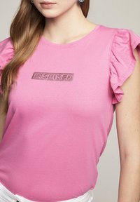 Vaaleanpunainen lyhythihainen t-paita, jossa on röyhelökaulukset ja "INSPIRED" -teksti kimaltavilla kirjaimilla suorakaiteen muotoisella paikkalla rinnassa. Pehmeä kankaan rakenne.