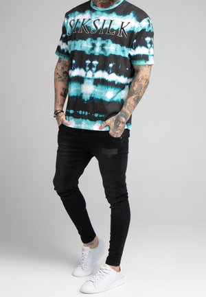 Camiseta de manga corta en patrón de tie-dye negro y verde azulado, con un logo de "SIKSILK". Combinada con jeans ajustados negros y zapatillas blancas.