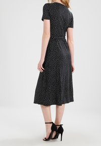 Czarna sukienka w kropki z krótkimi rękawami, podkreśloną talią i spódnicą o średniej długości. Noszona z czarnymi sandałami na wysokim obcasie. Gładka tekstura materiału.