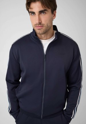 Mann trägt eine marineblaue Jacke mit Reißverschluss und weißen Streifen an den Ärmeln sowie ein weißes Hemd darunter, schaut leicht zur Seite.