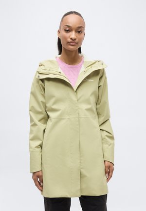 Mladá žena oblečená v svetlozelenej nepremokavej bunde s kapucňou cez ružové tričko, stojaca pred jednoduchým bielym pozadím.