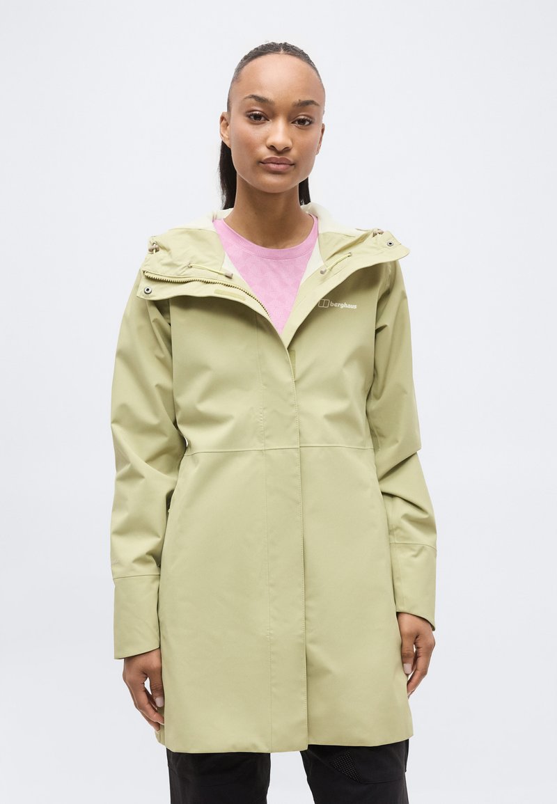 Νέα γυναίκα που φοράει μια ανοιχτοπράσινη αδιάβροχη κουκούλα πάνω από ένα ροζ τοπ, στέκεται μπροστά σε απλό λευκό φόντο.