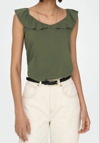 Top senza maniche color verde oliva con scollatura a volant, indossato con pantaloni di colore crema e una cintura nera. Tessuto morbido, design attillato.