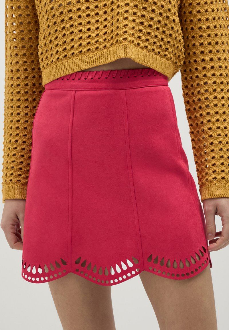 Primo piano di una persona che indossa una gonna rosa con tagli a goccia scallopati all'orlo e un maglione all'uncinetto giallo senape.