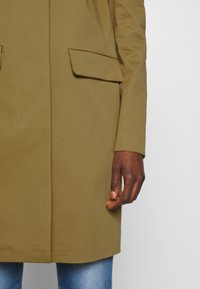 Main à la peau foncée à côté d'un manteau kaki avec poches à rabat, associée à un jean bleu clair sur un fond gris uni.