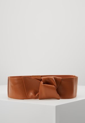 Brauner breiter Ledergürtel mit großem, verdrehtem Knotendetail, präsentiert auf einem weißen Podest vor einem schlichten Hintergrund.