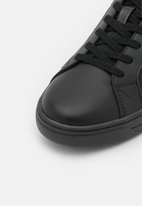 Gros plan sur le bout d'une basket noire avec des lacets noirs et une semelle en caoutchouc texturée sur fond blanc.