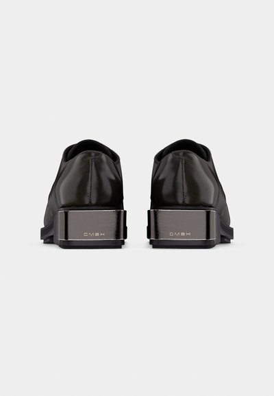 Λουστρινένια μαύρα παπούτσια σε φόρεμα, εμφανιζόμενα από πίσω, με χοντρούς μεταλλικούς τακούνια χαραγμένους με "GMBH" σε ένα απλό γκρι φόντο.