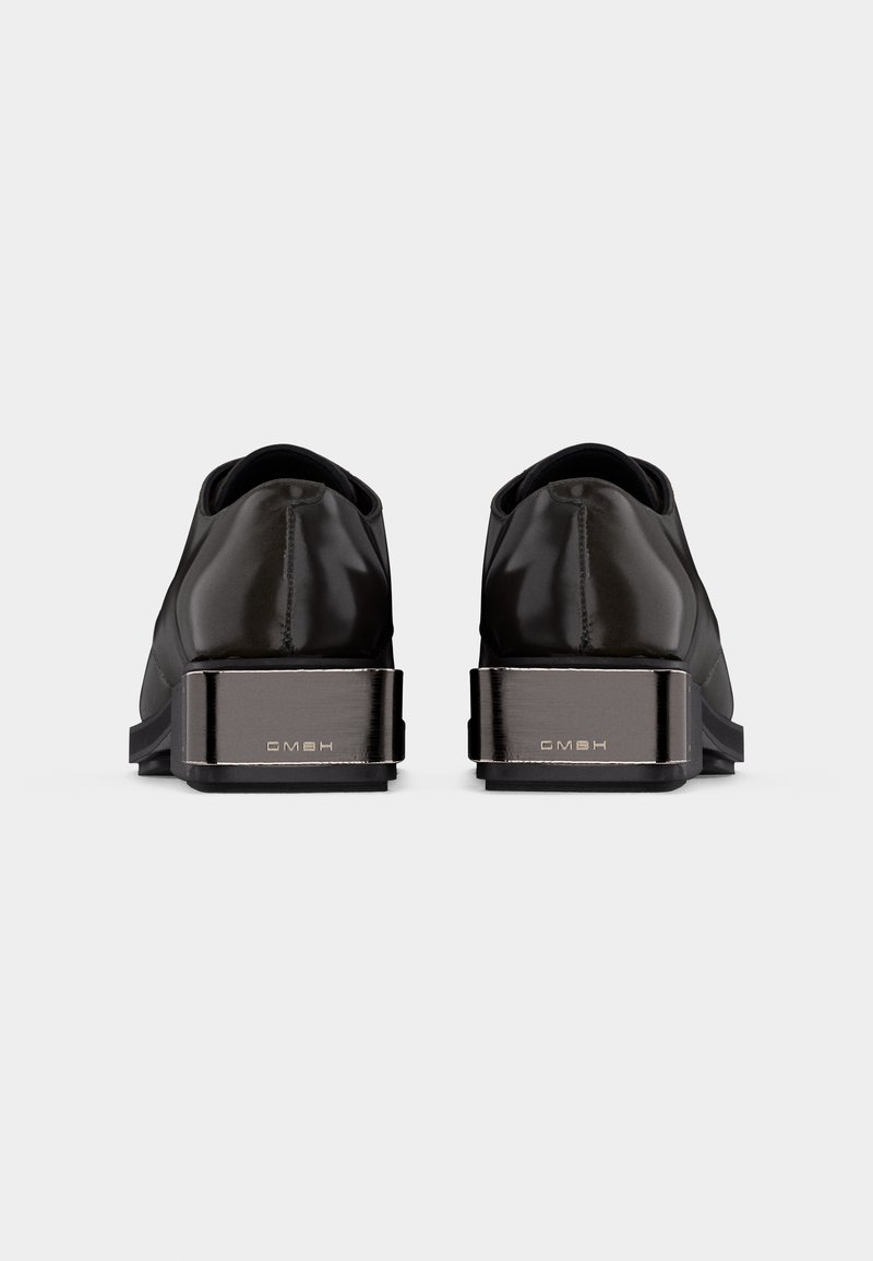 Shiny black dress shoes shown from the back, featuring thick metallic heels engraved with "GMBH" on a plain gray background.