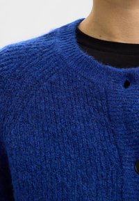 Niebieski dziergany kardigan z teksturowaną powierzchnią; posiada okrągły dekolt, zapięcie na guziki oraz mały otwór ozdobny w pobliżu kołnierza.