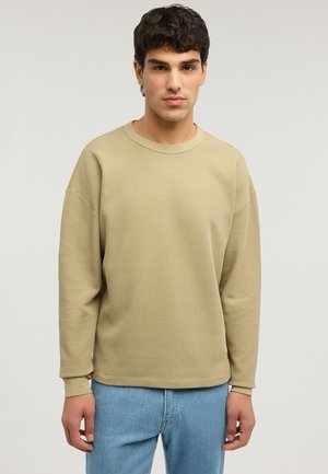 Junger Mann mit dunklem lockigem Haar, der ein beiges, strukturiertes Langarmshirt und hellblaue Jeans vor einem schlichten hellen Hintergrund trägt.