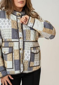 Jachetă din material patchwork, în culori estompate, cu diverse modele geometrice, butoane rotunde, guler înalt și două buzunare frontale cu margini îmbrăcate.