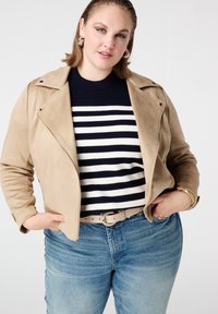 Beige jas van kunstsuède met ritssluiting, gedragen over een marineblauw-gestreepte trui en lichtblauwe spijkerbroek, met een gevlochten riem en gouden accenten.