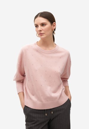 Suéter de punto rosa claro con hombros caídos, cuello redondo y adornos dispersos similares a gemas. Puños y dobladillo de canalé. Combinado con pantalones a rayas.