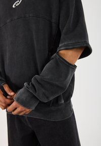 Svart hettegenser med vid halslinning og ribbestrikkede mansjetter. Har oppbrettede ermer som viser innervirkens struktur. Enkel logo på brystet.