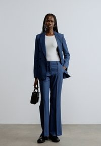 Modrý oblek s dvojradovým blejzrom, na mieru šité nohavice, nosené cez biely top. Modelka nesie malú čiernu kabelku a čierne topánky. Jednoduché pozadie.