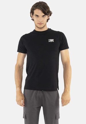 Jeune homme debout portant un t-shirt noir à manches courtes avec le logo "LEONE 1947" et un pantalon cargo gris sur un fond blanc uni.