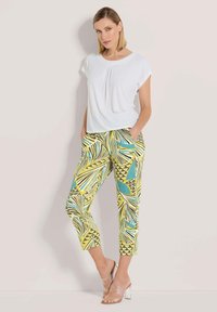 Weißes Kurzarmoberteil kombiniert mit einer Caprihose, die ein lebhaftes geometrisches Muster in Gelb, Blau und Schwarz aufweist. Transparente Absatzsandalen.