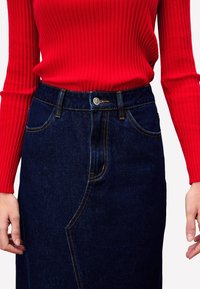 Jupe en denim bleu foncé avec des coutures visibles, poches avant et un bouton à la taille, associée à un haut rouge ajusté en ribbed.