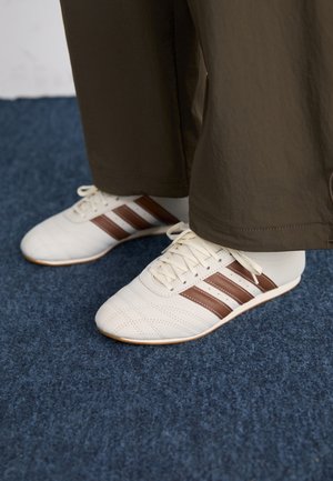 Hvide sneakers med brune striber båret med brune bukser på blåt tæppe, kun de nederste ben og fødder vist.