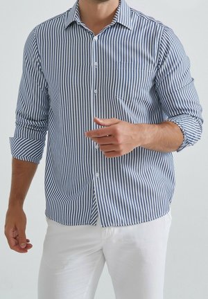 Chemise à manches longues rayée bleu et blanc avec un col boutonné et une poche poitrine, fabriquée dans un tissu léger. Présente des manches retroussées.