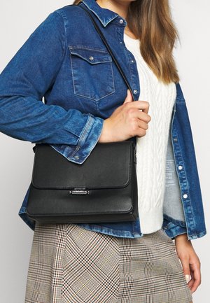 Kobieta w niebieskiej dżinsowej kurtce na białym swetrze w warkocze i spódnicy w kratę, trzymająca czarną skórzaną torebkę na ramię.