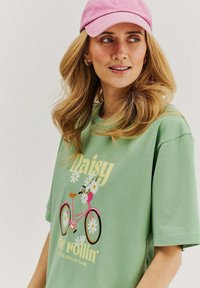 Woman with wavy blonde hair wearing a pink cap and light green T-shirt with a pink bicycle and daisy flower design.