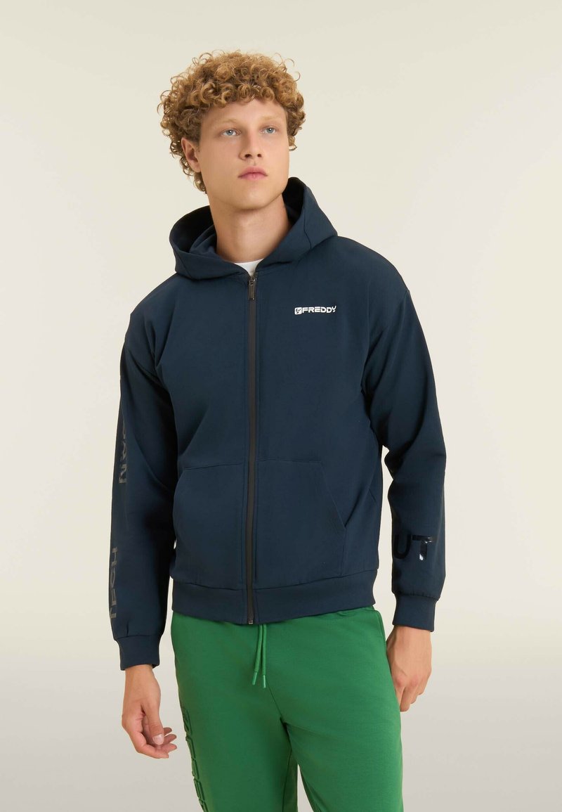 Giovane uomo che indossa una felpa blu navy con zip con il logo "Freddy" e pantaloni sportivi verdi, in piedi su uno sfondo chiaro e uniforme.